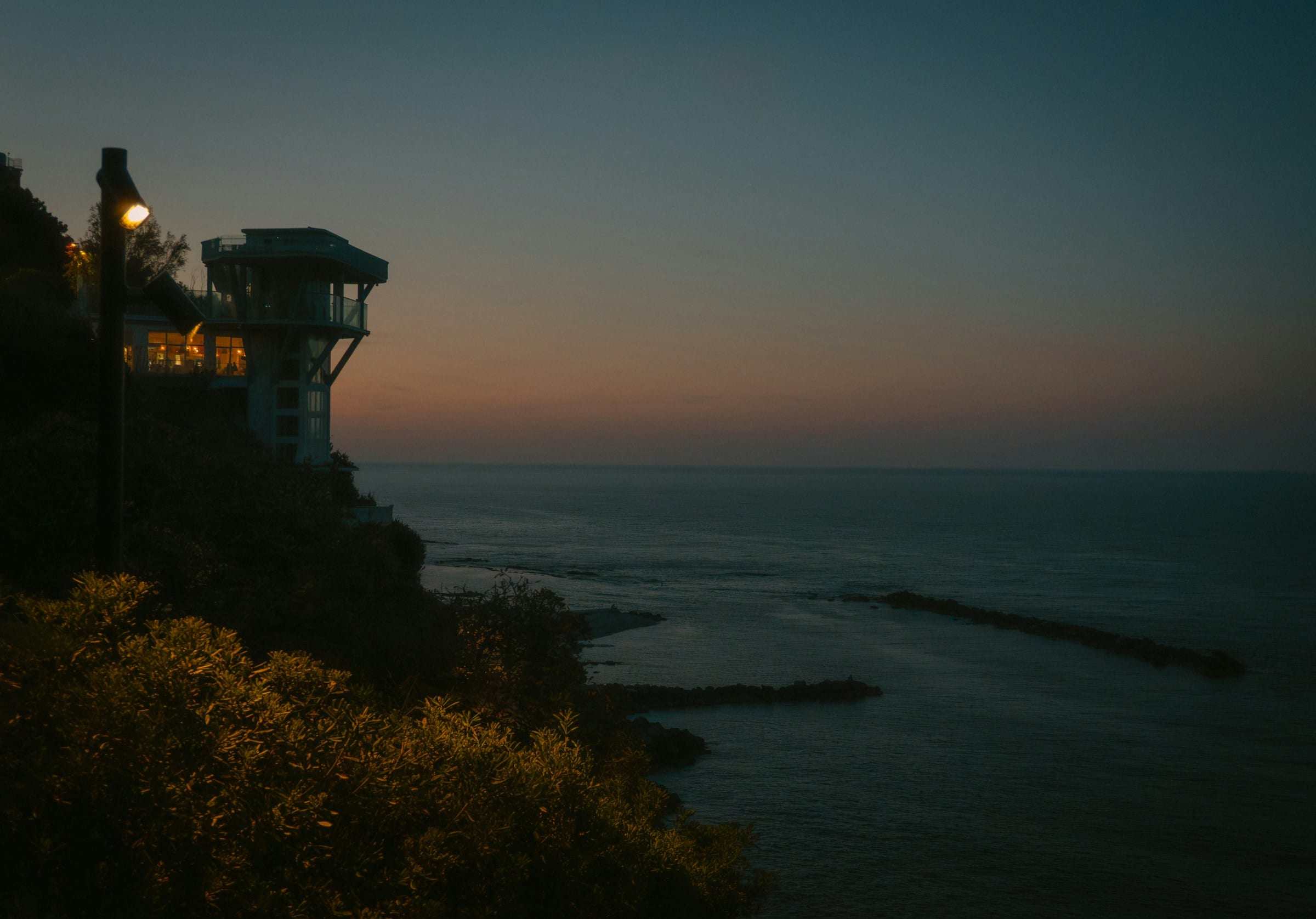 A coastal house on a headland at dusk, its lit windows standing over a calm sea.