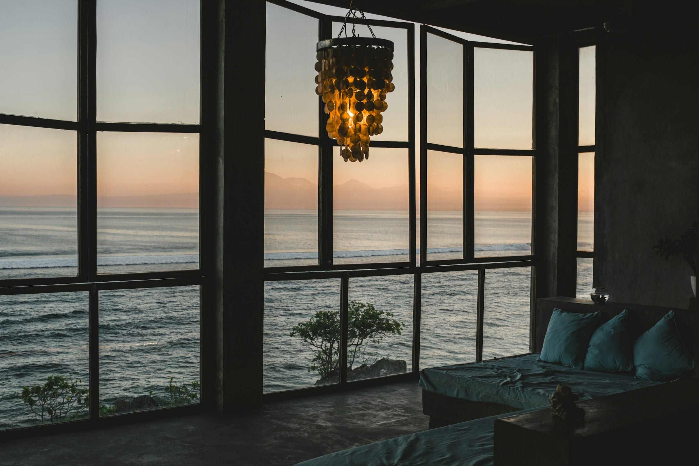 A room at sunset, floor to ceiling windows framing the sea, a shell chandelier over the glass.